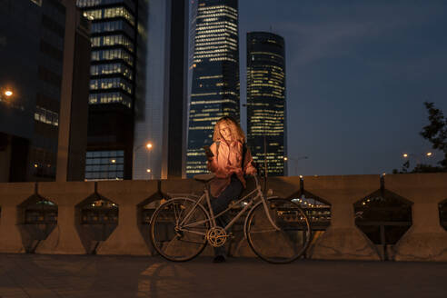 Junge Frau mit Fahrrad und Mobiltelefon in einer modernen Stadt bei Nacht - JCCMF07691