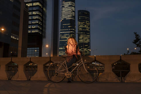 Young woman with bicycle using mobile phone in modern city at night - JCCMF07691
