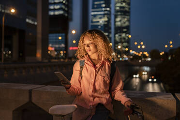 Woman with curly hair using mobile phone in city at night - JCCMF07692
