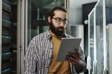 Technician wearing eyeglasses looking at tablet PC in server room - DSHF00578