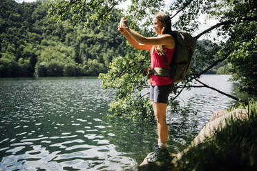 Smiling woman photographing lake through smart phone - GIOF15573