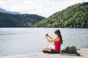 Frau fotografiert den Levico-See mit dem Smartphone auf dem Steg sitzend - GIOF15598