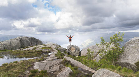 Young hiker with arms raised standing on rock under cloudy sky - JAQF01079