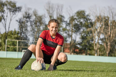Happy girl with soccer ball crouching on field on sunny day - ZEDF04911