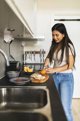 Smiling young woman cutting orange fruit in kitchen - DMMF00212