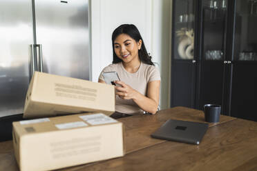 Smiling woman checking home delivery box at dining table - DMMF00229