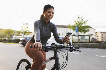Smiling woman using mobile phone sitting on bicycle at street - DMMF00249