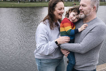 Glückliche Eltern mit Tochter stehen vor einem See im Park - AMWF00841