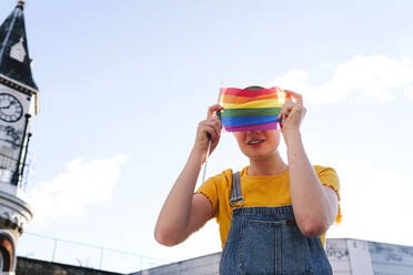 Lächelnde nicht-binäre Person, die ihr Gesicht mit einer Regenbogenflagge bedeckt - ASGF03003