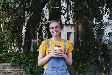 Happy non-binary person with disposable coffee cup in front of plants - ASGF03007