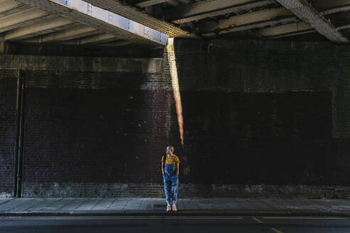 Young androgynous person waiting on sidewalk below bridge - ASGF03010