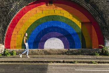 Young androgynous person walking on footpath by rainbow wall - ASGF03014