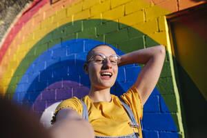 Glückliche androgyne Person mit Hand hinter rasiertem Kopf, die ein Selfie vor einer Regenbogenwand macht - ASGF03018