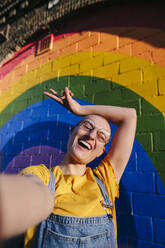 Fröhliche Transgender-Person, die ein Friedenszeichen macht und ein Selfie vor einer Regenbogenwand schießt - ASGF03019