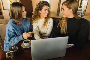 Fröhliche Freundinnen, die mit Laptop und Smartphone am Tisch sitzen, heißen Tee trinken und miteinander kommunizieren - ADSF39525