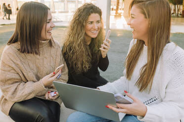 Gruppe von glücklichen Freundinnen in legerer Kleidung auf einer Steinbank auf dem Campus sitzend, während sie gemeinsam auf Laptop und Smartphones surfen - ADSF39530