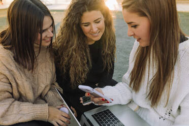 Group of happy female friends in casual outfits sitting on stone bench in campus while browsing on laptop and smartphones together - ADSF39531