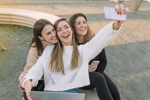 Hoher Winkel von fröhlichen jungen Freundinnen, die ein Selfie mit ihrem Smartphone machen und auf einer Steinbank in der Stadt sitzen - ADSF39532