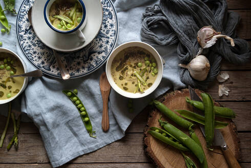 Flache Schalen mit grüner Erbsen- und Kokosnusscremesuppe auf Holztisch mit Erbsenschoten und Knoblauch in Komposition - ADSF39541