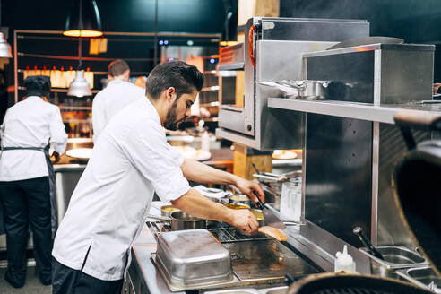 Seitenansicht eines männlichen Kochs mit Mütze und Schürze, der in einer modernen Restaurantküche Lachs zubereitet - ADSF39562