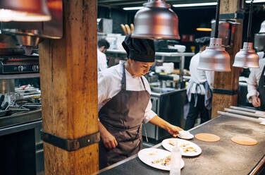 Adult woman in apron and hat preparing and serving dishes on kitchen in modern restaurant - ADSF39564