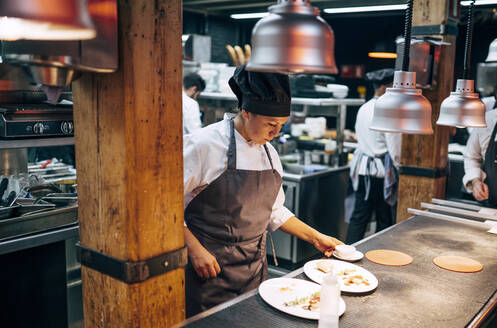 Erwachsene Frau in Schürze und Hut bei der Zubereitung und dem Servieren von Speisen in der Küche eines modernen Restaurants - ADSF39564