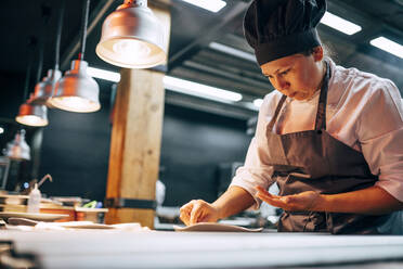 Erwachsene Frau in Schürze und Hut bei der Zubereitung und dem Servieren von Speisen in der Küche eines modernen Restaurants - ADSF39565