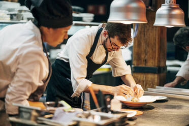 Man and woman in hats and aprons working in team and serving dishes with delicious food in kitchen - ADSF39568