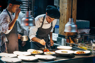 Men in hats and aprons working in team and serving dishes with delicious food in kitchen - ADSF39570