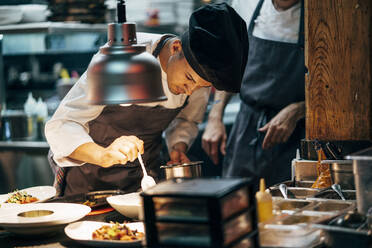 Men in hats and aprons working in team and serving dishes with delicious food in kitchen - ADSF39571