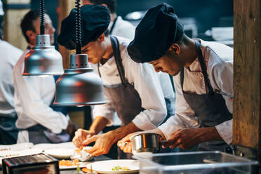 Men in hats and aprons working in team and serving dishes with delicious food in kitchen - ADSF39573