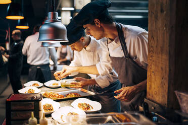 Men in hats and aprons working in team and serving dishes with delicious food in kitchen - ADSF39577