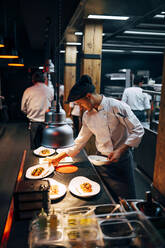 Side view of professional chef in hat serving similar dishes in row working on kitchen of modern restaurant - ADSF39578