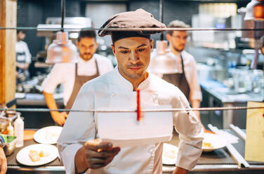 Chef in hat reading order on paper hanging on string while working in modern kitchen of restaurant - ADSF39581