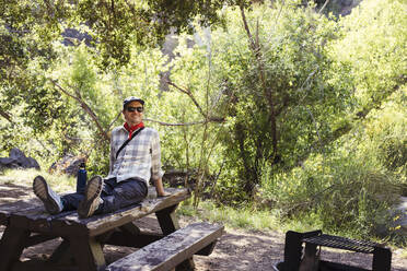 Happy man wearing sunglasses on wooden table in forest - ACTF00260