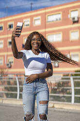 Happy young woman with tousled hair taking selfie through smart phone on footpath - OCMF02546