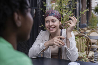 Glückliche Frau mit Kopftuch im Gespräch mit einem Freund in einem Straßencafé - AMWF00855