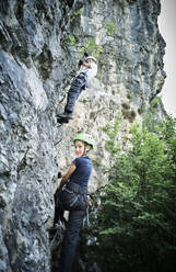 Siblings climbing rocky cliff in forest - DIKF00735