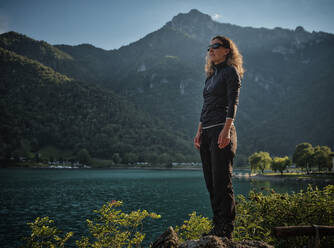 Ältere Frau mit Sonnenbrille steht auf einem Felsen am See - DIKF00765