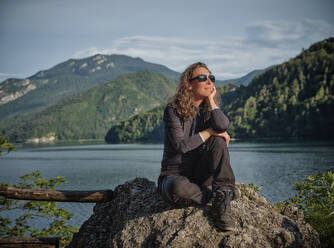 Smiling thoughtful woman sitting on rock in front of lake - DIKF00766