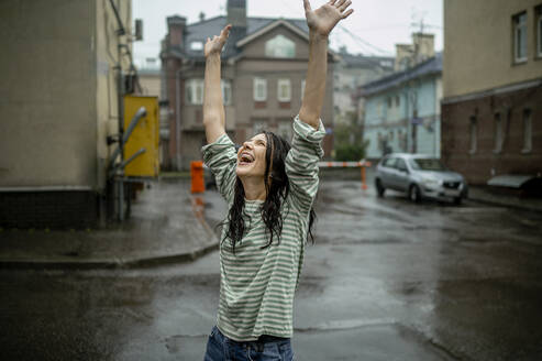 Glückliche Frau mit erhobenen Armen genießt den Regen in der Stadt - ANAF00190