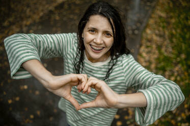 Happy woman gesturing heart with hands in park - ANAF00191