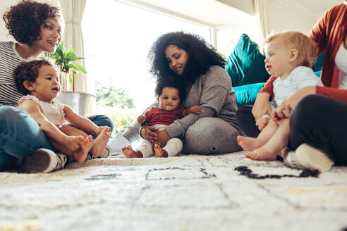 Fröhliche multiethnische Freunde mit Kindern, die zu Hause sitzen. Mütter im Wohnzimmer schauen ihren Babys beim Spielen zu - JLPSF10397