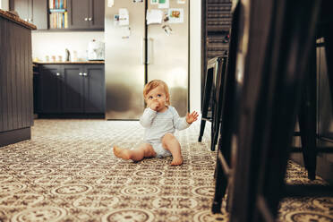 Baby sitting on floor eating something. Cute child in kitchen at home. - JLPSF10404