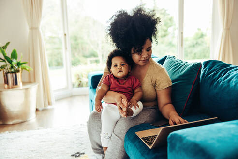 Afroamerikanische Frau mit Baby auf dem Sofa sitzend und Laptop benutzend. Freiberuflerin arbeitet während des Mutterschaftsurlaubs. - JLPSF10406