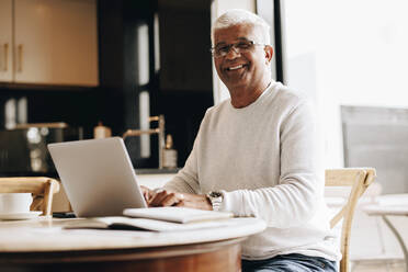 Glücklicher älterer Geschäftsmann, der in die Kamera lächelt, während er mit einem Laptop in seinem Büro zu Hause sitzt. Älterer Geschäftsmann, der eine geschäftliche E-Mail tippt, während er von zu Hause aus arbeitet. - JLPSF10411