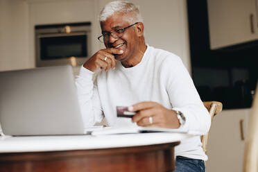 Unbekümmerter älterer Mann, der fröhlich lächelt, während er zu Hause online einkauft. Glücklicher reifer Mann, der seine Kreditkarte benutzt, um eine Online-Bestellung auf einem Laptop aufzugeben. Rentner, der sich nach seiner Pensionierung verwöhnen lässt. - JLPSF10425