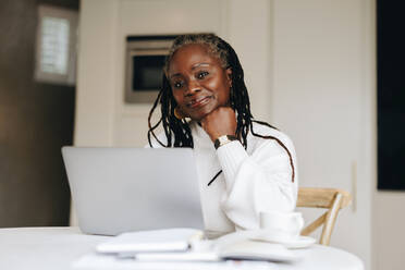Ältere Geschäftsfrau, die in die Kamera schaut, während sie zu Hause an einem Laptop arbeitet. Ältere Geschäftsfrau, die in ihrem Büro zu Hause freiberuflich arbeitet. - JLPSF10442