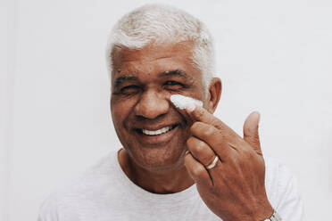 Happy senior man smiling at the camera while applying a moisturizing cream on his face. Body positive senior man treating his aging skin with a nourishing beauty product. - JLPSF10465