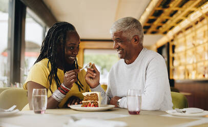 Ein glückliches älteres Paar, das sich anlächelt, während es in einem Café einen leckeren Kuchen isst. Ein sorgloses älteres Paar, das sich in einem Restaurant amüsiert. Ein reifes Paar, das seinen Ruhestand gemeinsam genießt. - JLPSF10468